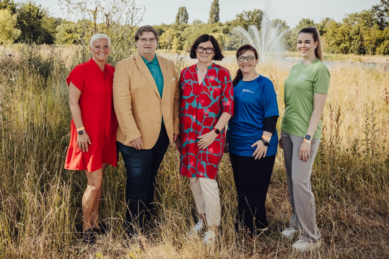 Fünf Personen posieren lachend für ein Gruppenfoto vor einer natürlichen Kulisse mit Wiese