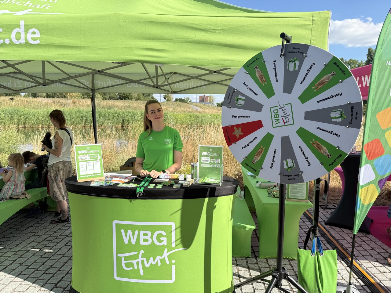 Mitarbeiterin der WBG Erfurt steht hinter einem Infostand mit Glücksrad, umgeben von Flyern und Werbegeschenken