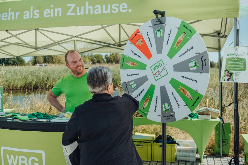 Tag der Genossenschaft WBG Erfurt Person dreht an einem Glücksrad mit verschiedenen Symbolen und Texten vor einem Stand mit grünem WBG-Logo