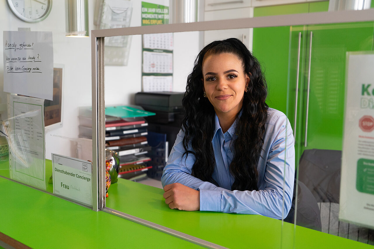 Concierge-Mitarbeiterin hinter einer grünen Empfangstheke mit Schutzscheibe im Servicebereich eines Wohngebäudes.