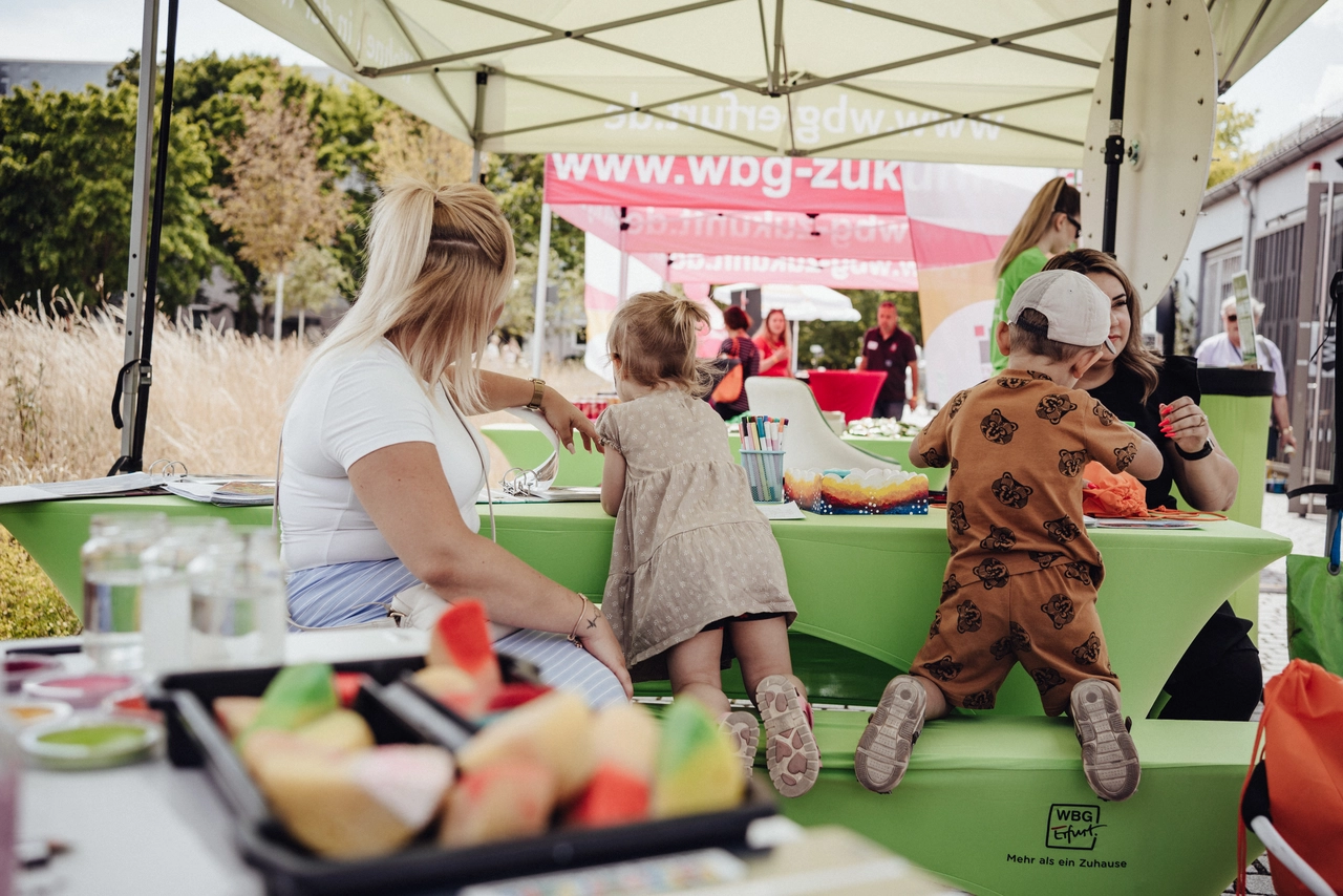 Kinder und Eltern sitzen unter einem Pavillon an einem Basteltisch und gestalten bunte Kunstwerke